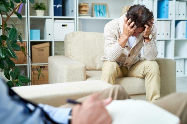Understanding Persistent Depressive Disorder (Dysthymia): Symptoms, Causes, and Treatment. Depressed emotional young man sitting in armchair and holding head in hands while crying at psychotherapy session. - East Coast Telepsychiatry Understanding Persistent Depressive Disorder (Dysthymia): Symptoms, Causes, and Treatment. Depressed emotional young man sitting in armchair and holding head in hands while crying at psychotherapy session.